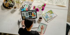 A graphic designer sits at her colorfully cluttered desk using a tablet and a laptop